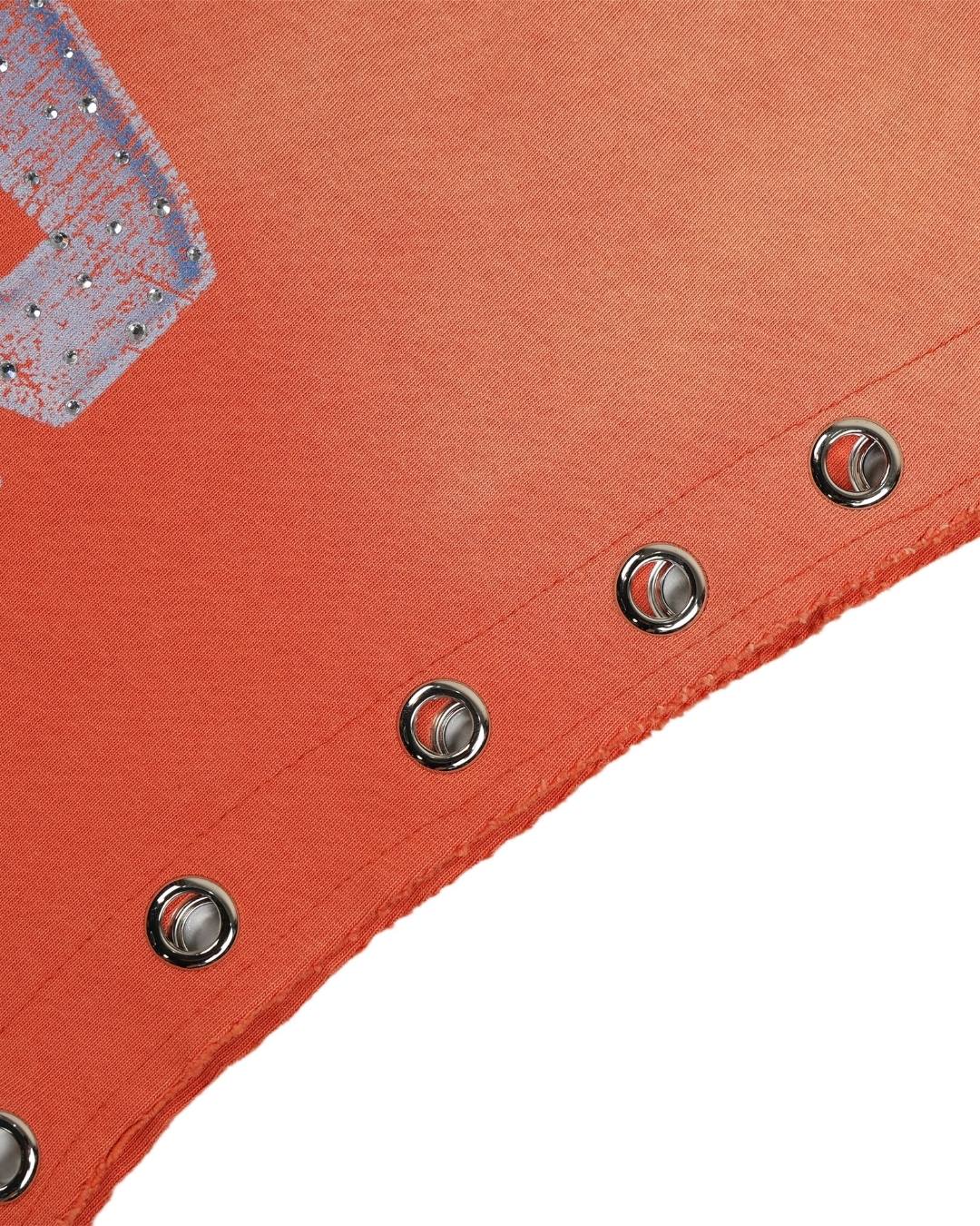 Close-up of a textured orange fabric product, likely a belt or strap, featuring a diagonal row of silver metal grommets and a glittery silver graphic detail on the upper left, set against a plain white background.